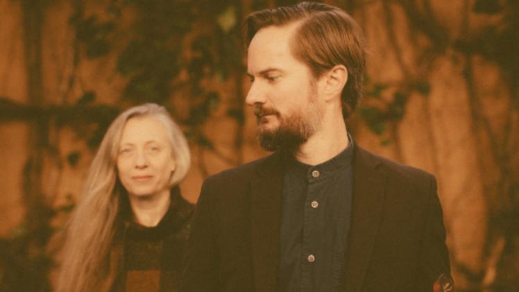 Two people standing, none person standing in front holding a guitar and looking behind at other person who is looking directly at the camera.